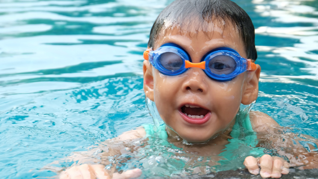 Young boy wearing goggles, holding on to the side of an indoor swimming pool