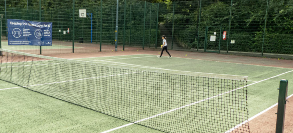 Person at the far end of a tarmac tennis court, bouncing a tennis ball